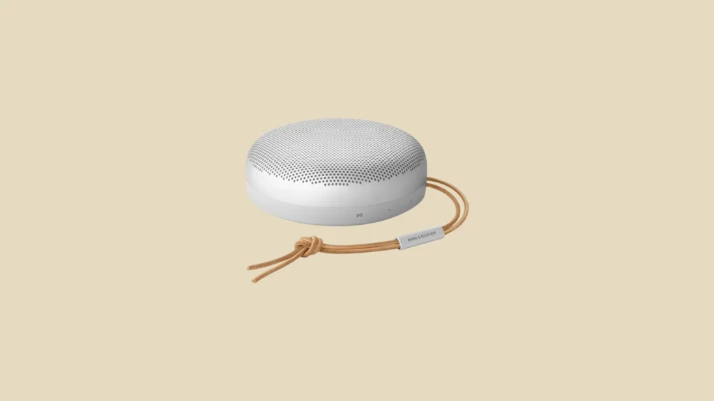 Front view of a round, white Bluetooth speaker with perforated top and brown leather strap on a beige background.