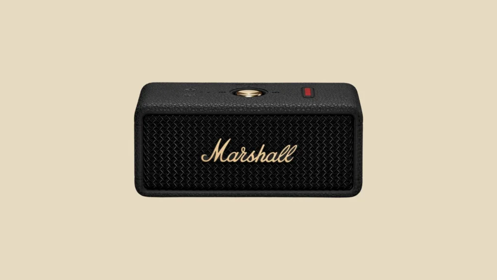 Front view of black Marshall Bluetooth speaker with gold logo, top gold button, and red indicator on beige background.