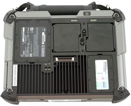 Rear view of a rugged laptop underside with compartments, battery slots, vents, ports, labels, and corner bumpers. Brand/model unspecified.