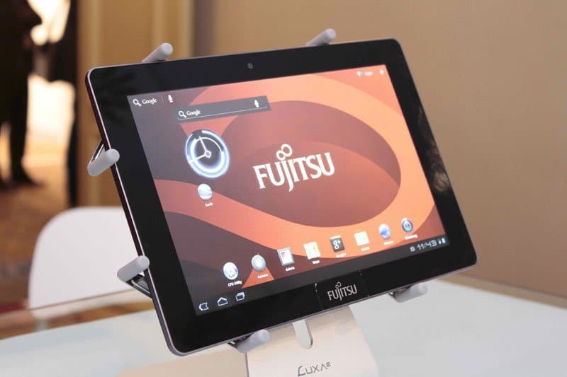 Front view of a Fujitsu tablet on a stand in landscape, home screen visible with Fujitsu logo and app icons.