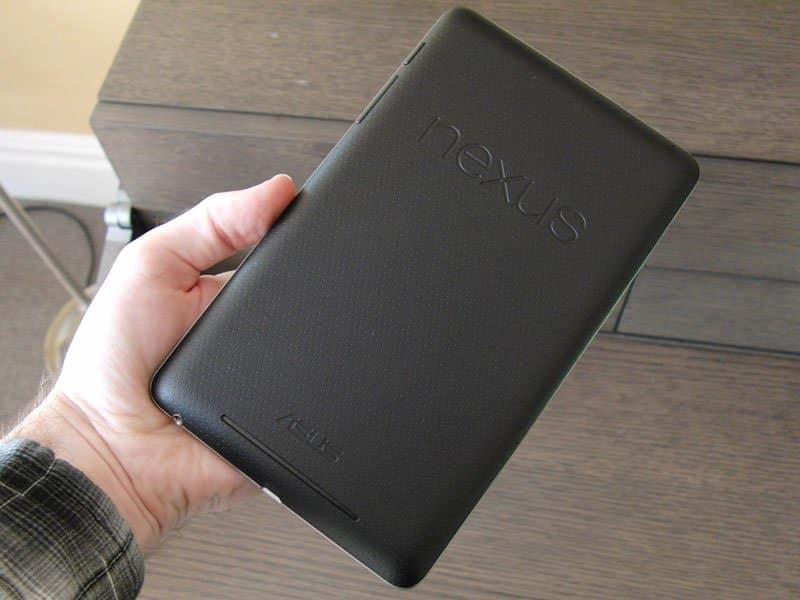 Rear view of a person holding a black Google Nexus tablet, showing the embossed "nexus" logo against a wooden background.