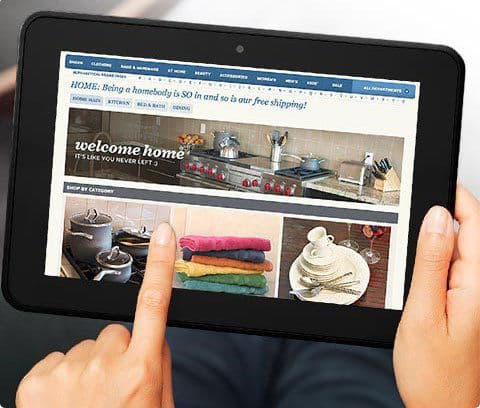 Person holding a front-facing Apple iPad showing a home goods website; finger points at kitchen, dishes, and towels categories.