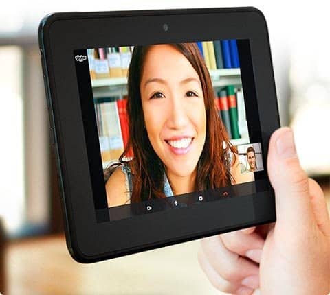 Front view of a person holding a tablet on a video call; main screen shows smiling woman, bookshelves behind her.