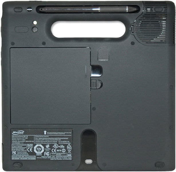 Rear view of rugged black tablet with top handle, right speaker, large battery panel, ports, screws, and regulatory labels.