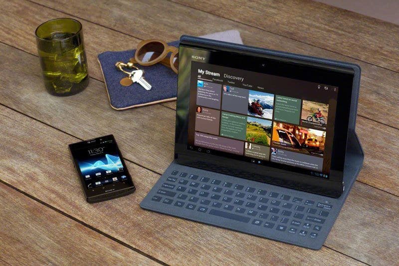 Front view of an iPad Pro with keyboard case showing news feed, next to smartphone, drink, sunglasses, and keys on wooden table.