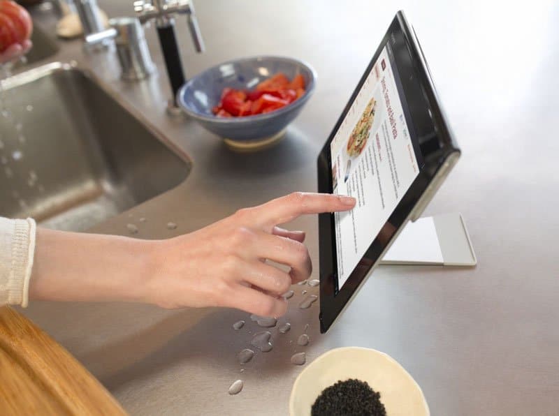 Front view of a tablet on a wet kitchen counter, recipe on screen, hand touching display, food bowls nearby. Brand/model unclear.