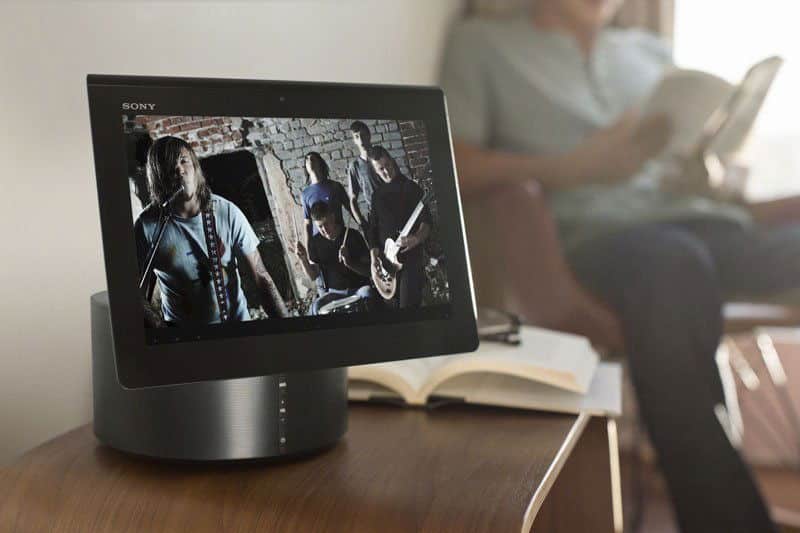 Front view of Sony smart display on table streaming music video; background shows person reading with notebook nearby.