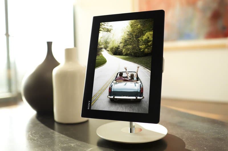 Front view of a digital photo frame on table showing two people in a convertible; vases and blurred painting in background.