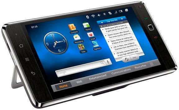 Black touchscreen device in landscape, front view; blue home screen with clock, apps, news feed; built-in kickstand deployed.