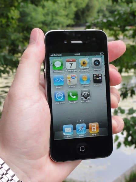 Front view of a black Apple iPhone 4 held in hand, showing home screen app icons; green trees blurred in the background.
