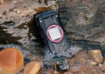 Casio GzOne Ravine front view, black rugged phone with pink circular display, resting on rocks in shallow water