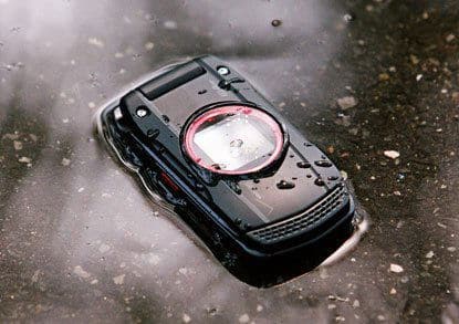 Casio GzOne Ravine phone, front view, with large circular dial on dark rugged casing, sitting on wet surface with water droplets