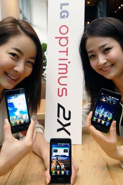 Two women holding LG Optimus 2X smartphones (front view) with a product sign between them on a wooden table indoors.