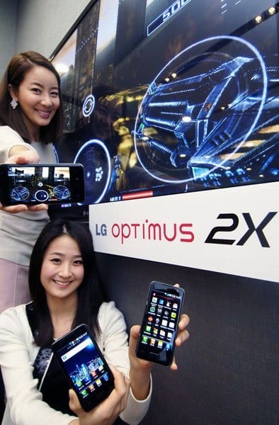 Two women hold LG Optimus 2X smartphones (front view) in front of a large screen showing a car dashboard interface.