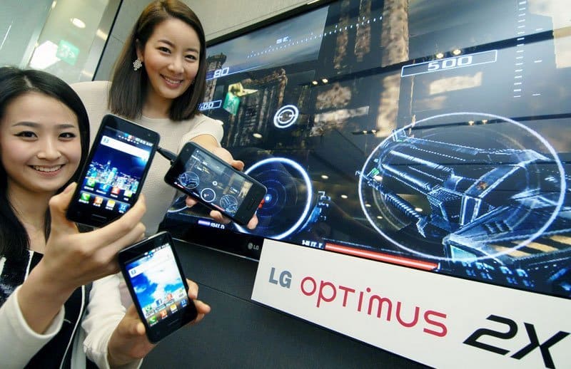 Two women hold LG Optimus 2X phones (front view) in front of a video game screen; logo appears prominently in foreground.