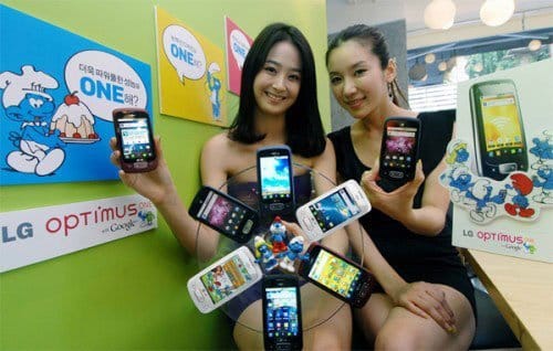 Two women displaying multiple LG Optimus One smartphones arranged in a circle, with LG branding visible on the wall behind them