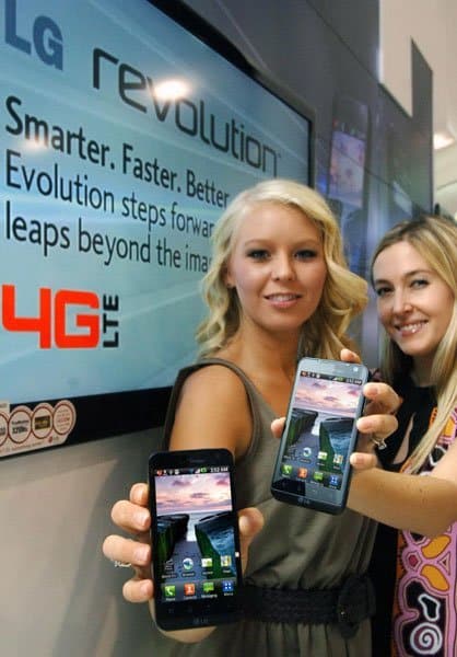 Two women display LG Revolution smartphones front view, showing the home screen in front of a "LG Revolution 4G LTE" sign.