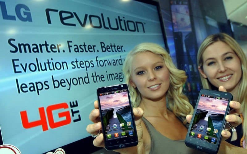 Front view of two women holding LG Revolution smartphones with home screens visible, standing before an LG 4G LTE promo display.