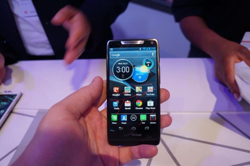 Front view of Motorola DROID RAZR HD in hand, home screen visible; two people seated at a table in the background.