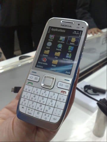 Front view of a Nokia E55 with physical keypad and color screen showing Applications menu, held in hand, connected to a black cable.