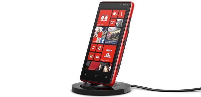 Nokia Lumia 820 front view with red case on charging dock, displaying Windows Phone home screen with app tiles
