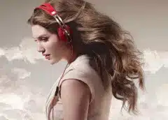 Woman in profile wearing over-ear headphones with pink accents in her hair, pictured against a cloudy sky background