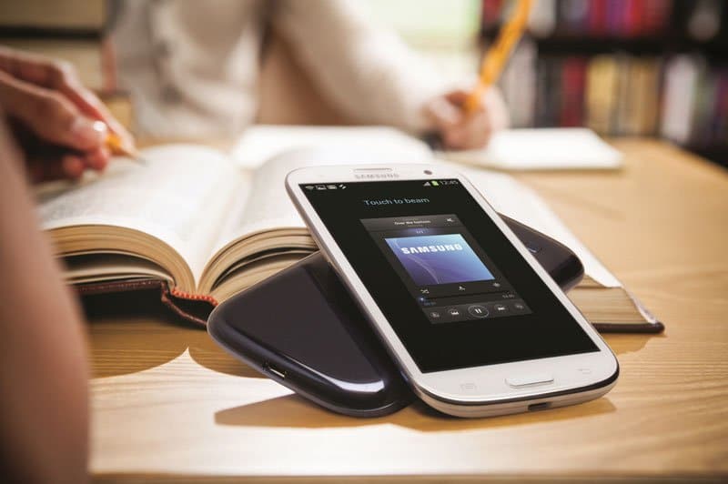 Samsung Galaxy S III smartphone displaying home screen, positioned on wooden desk next to open book and person holding pen