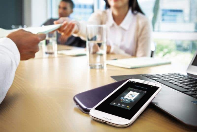 Samsung Galaxy S III on table during business meeting, screen-on view with colleagues and laptop in background