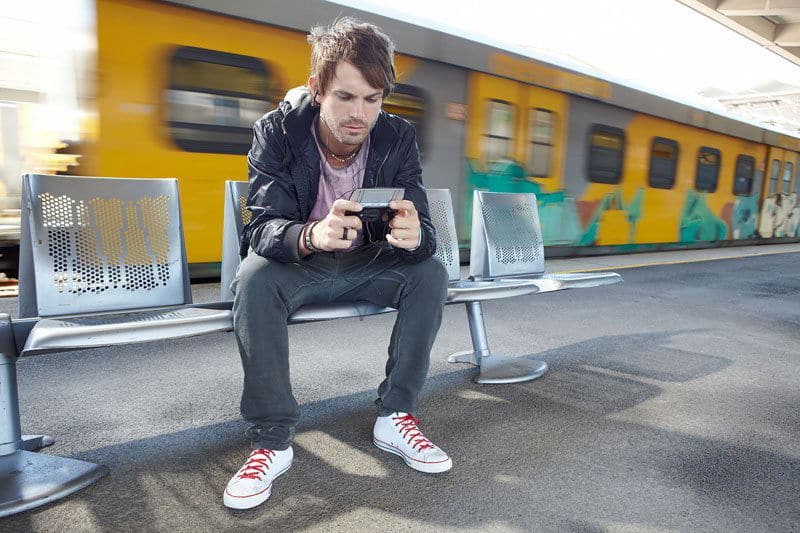 Man sitting on a bench at a train station, holding a Sony Ericsson Xperia Play gaming device with both hands