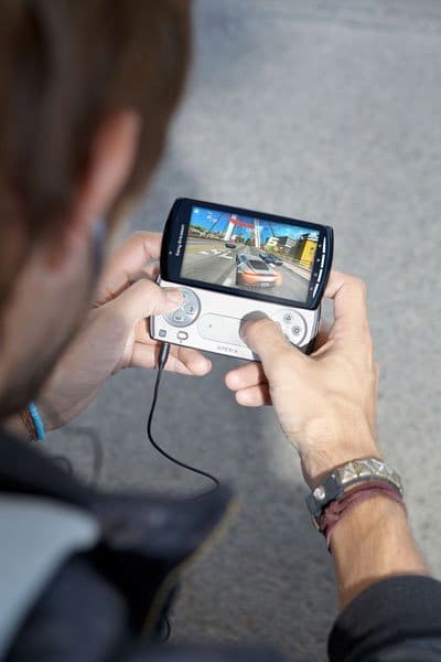 Sony Ericsson Xperia Play held in hands, displaying a racing game on screen with game controller buttons visible below the display