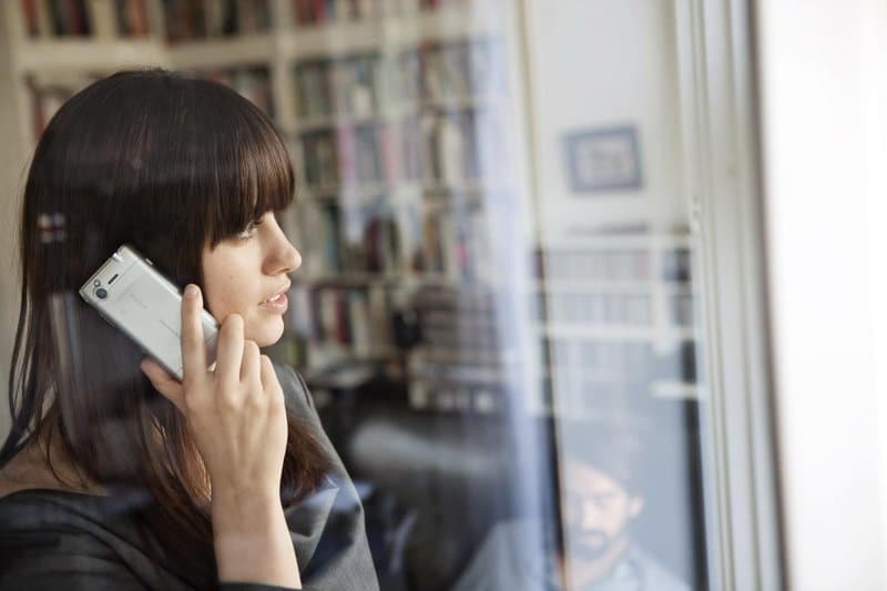 Woman holding Sony Ericsson Xperia X2 to her ear while looking out a window, with bookshelves visible behind her