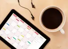 Ipad displaying a calendar app with month view, alongside a cup of coffee and earbuds on a tan surface