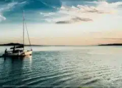 Sailboat floating on calm turquoise water at sunset with soft clouds and distant shoreline